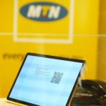 A tablet displaying a QR code, with the MTN logo in the background on a yellow backdrop.