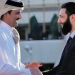Two men in formal attire shake hands with smiles, set against an outdoor background, depicting a friendly diplomatic encounter.
