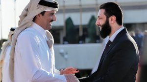 Two men in formal attire shake hands with smiles, set against an outdoor background, depicting a friendly diplomatic encounter.